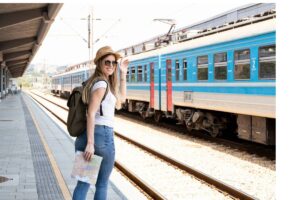 A woman with a backpack and a map waits at a train station, with a blue train visible on the tracks behind her.