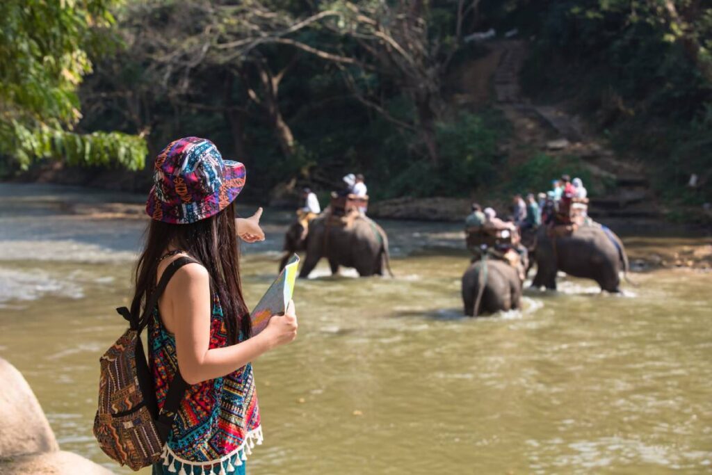 A woman in colorful attire points towards elephants crossing a river as tourists ride on their backs in a lush, green landscape.