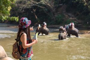 A woman in colorful attire points towards elephants crossing a river as tourists ride on their backs in a lush, green landscape.