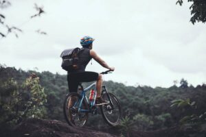 A cyclist in a blue helmet and backpack gazes at a lush forested landscape while perched on a rocky outcrop.