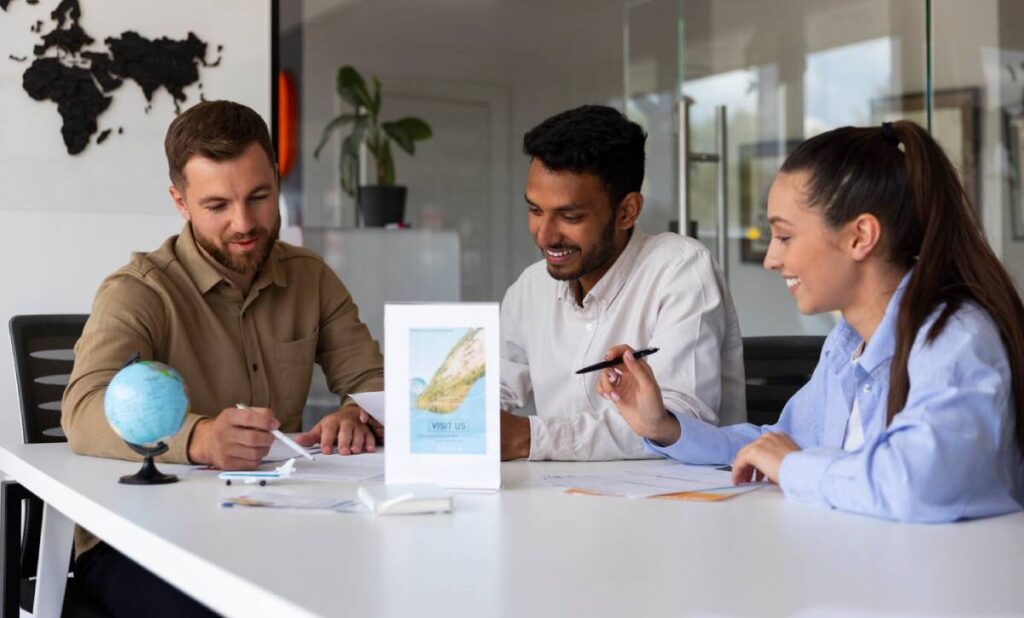 Three individuals engage in a collaborative meeting at a modern table, surrounded by papers, a globe, and a promotional sign.