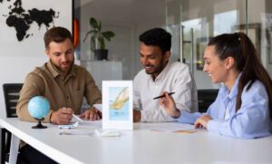 Three individuals engage in a collaborative meeting at a modern table, surrounded by papers, a globe, and a promotional sign.