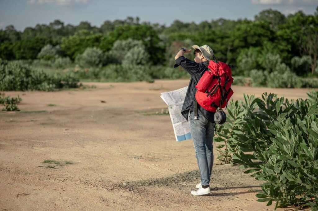A person with a red backpack stands on dry ground, holding a map while surveying the lush green landscape ahead.