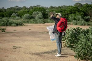 A person with a red backpack stands on dry ground, holding a map while surveying the lush green landscape ahead.