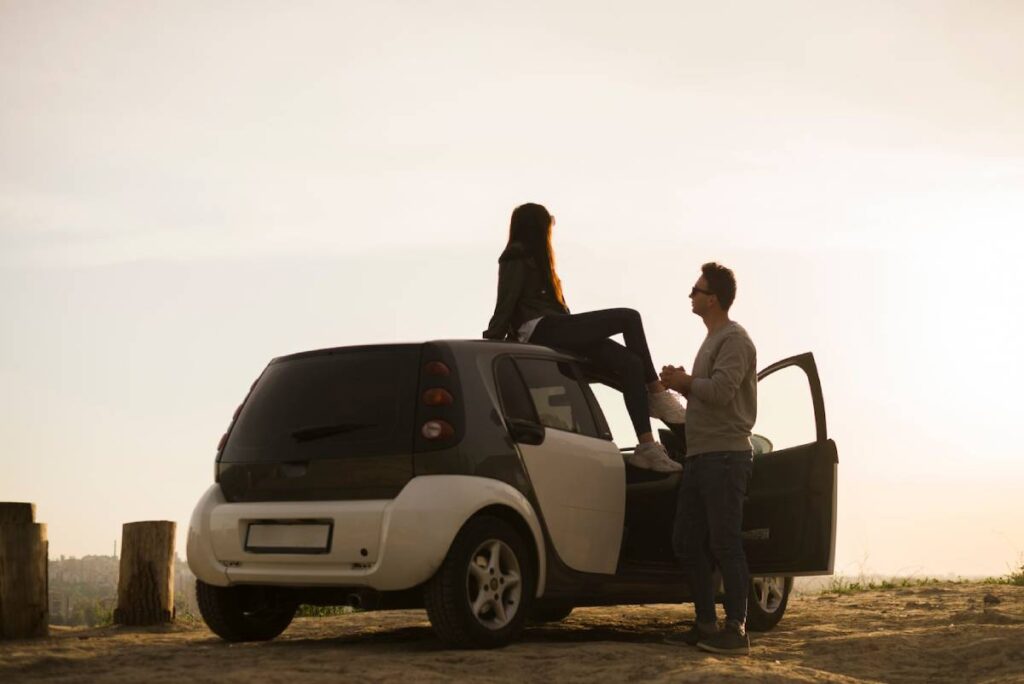 A couple enjoys a romantic moment beside a compact car, with one person sitting on the roof as the sun sets in the background.