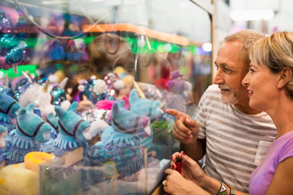 A couple gazes excitedly at colorful plush toys inside a claw machine, with vibrant lights reflecting in the glass.