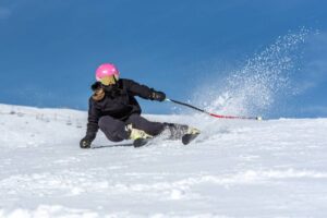 A skier in a pink helmet and black jacket carves through fresh snow against a clear blue sky, sending up a spray of powder.