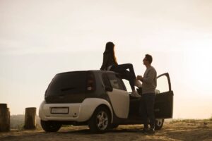 A couple enjoys a romantic moment beside a compact car, with one person sitting on the roof as the sun sets in the background.