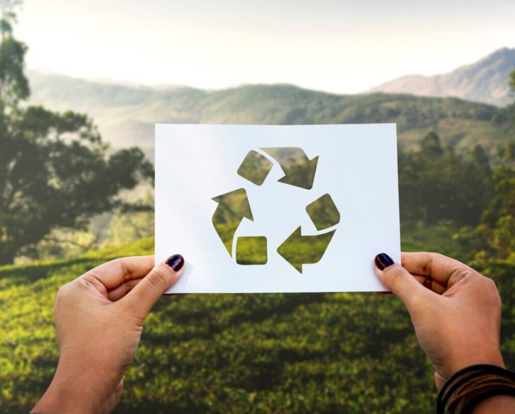Hands holding a white paper with a cut-out recycling symbol, set against a lush green landscape and mountains in the background.