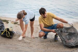 Two individuals collect litter on a beach, using gloves and bags to clean up debris near calm water.