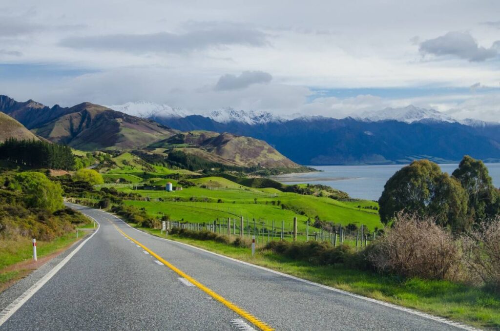 A winding road leads through lush green hills, with snow-capped mountains and a serene lake in the background, under a cloudy sky.