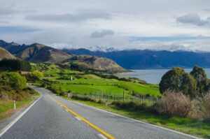 A winding road leads through lush green hills, with snow-capped mountains and a serene lake in the background, under a cloudy sky.