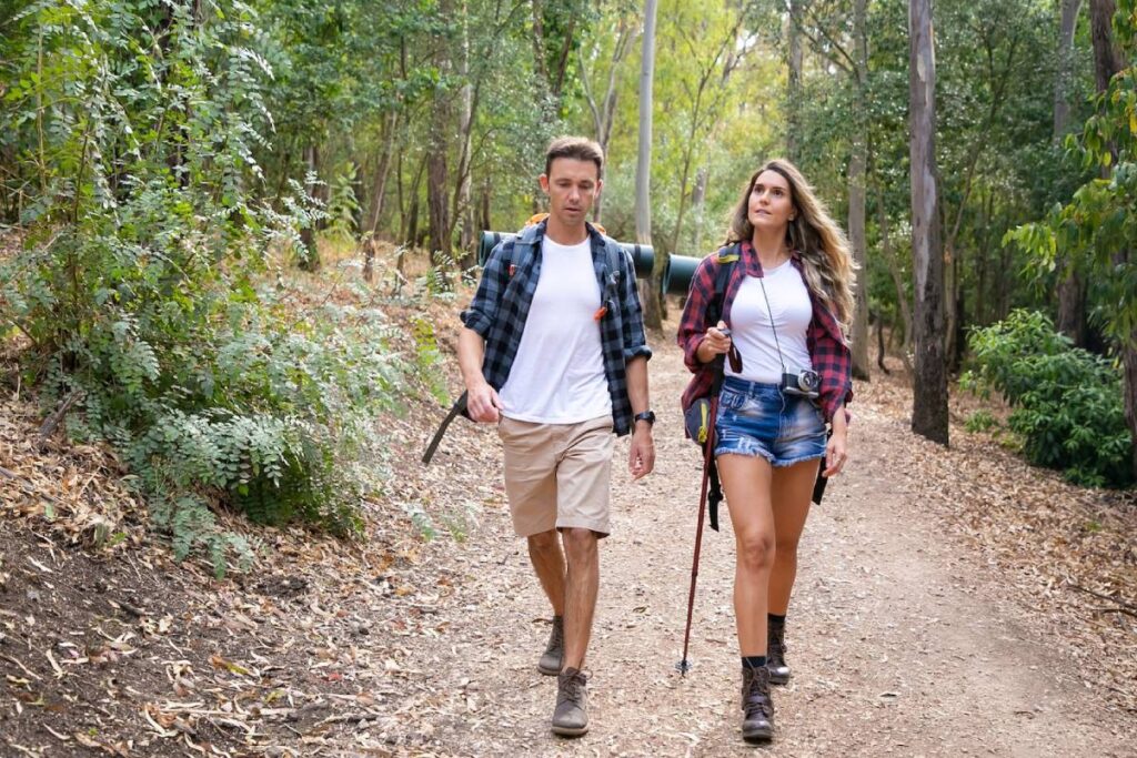 A couple walks hand in hand along a forest trail, dressed in casual hiking attire, surrounded by lush greenery and earthy textures.