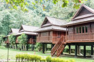 Four traditional wooden houses on stilts surrounded by lush greenery, set against a backdrop of dense trees and natural beauty.