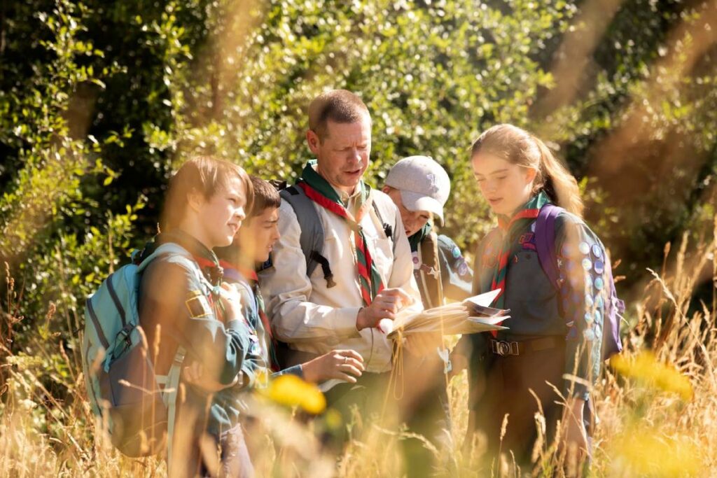 A group of young scouts gathers outdoors, examining a map together amidst tall grass and lush greenery, focused on their adventure.