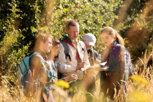 A group of young scouts gathers outdoors, examining a map together amidst tall grass and lush greenery, focused on their adventure.