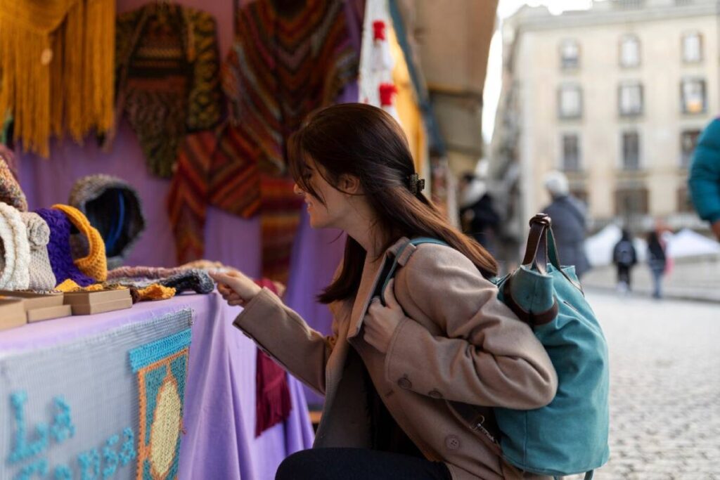 A woman examines handmade scarves and hats displayed on a purple table at a market, with colorful textiles hanging in the background.