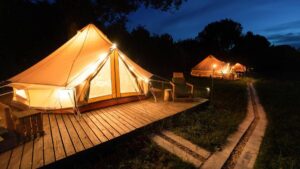Illuminated glamping tents on wooden decks under a twilight sky, with a winding path leading through lush grass.