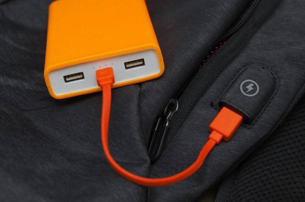 An orange power bank connected to a black jacket through an orange charging cable, with a zipper and a charging symbol visible.