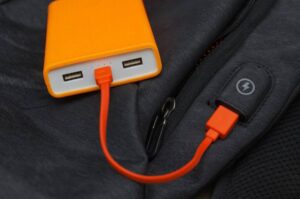 An orange power bank connected to a black jacket through an orange charging cable, with a zipper and a charging symbol visible.