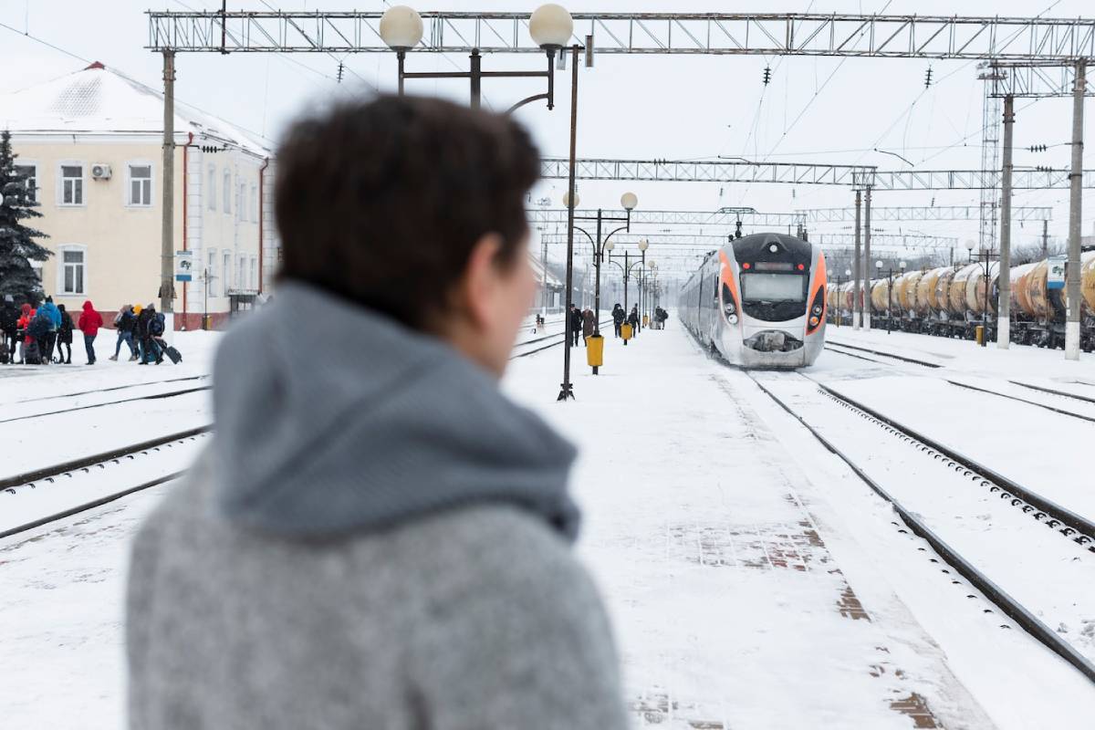 A snowy train station scene with a sleek train approaching, passengers gathering, and a historic building in the background. 