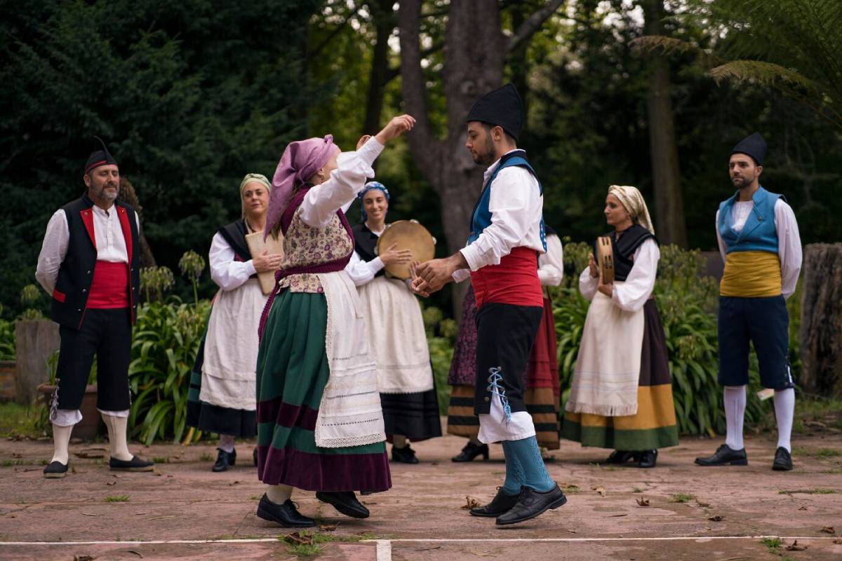 A group of people dressed in traditional attire perform a folk dance outdoors, surrounded by greenery and rustic scenery. 