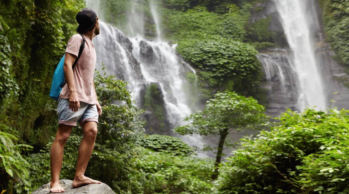  A young man with a backpack stands on a rock, gazing at a cascading waterfall surrounded by lush greenery.