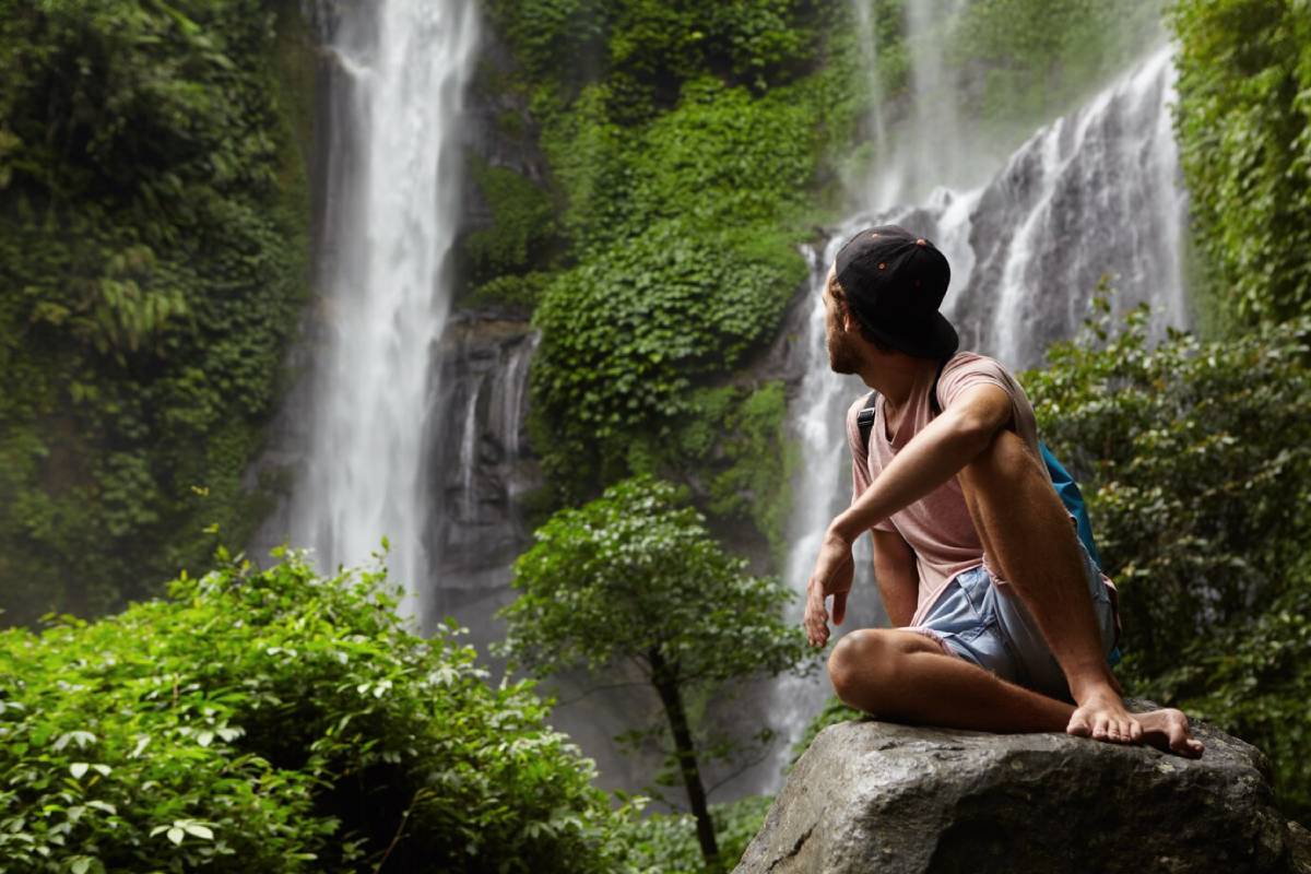  A man sits on a rock in front of a lush waterfall, gazing at the cascading water surrounded by green foliage.