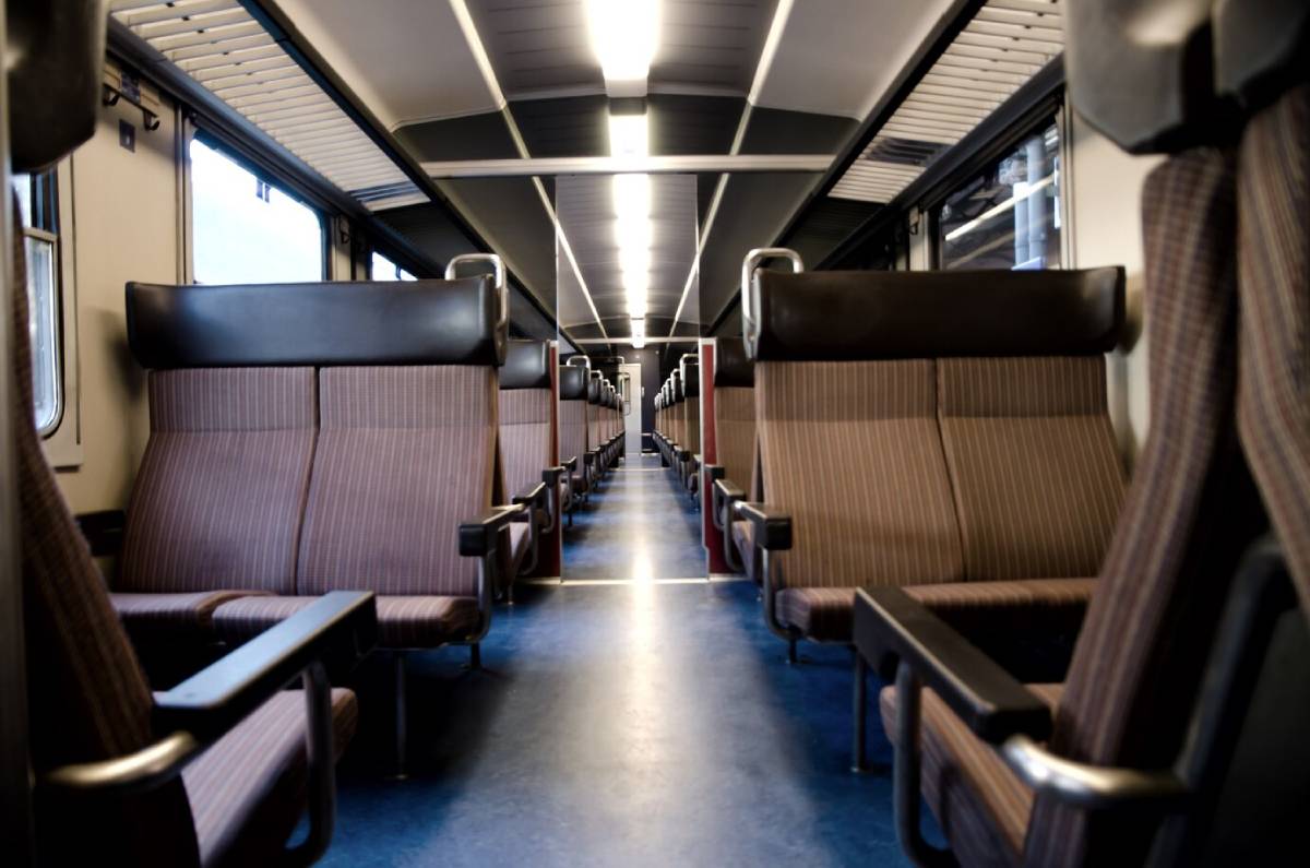  Interior of a train carriage featuring rows of empty, cushioned seats with armrests, and bright overhead lights illuminating the aisle.