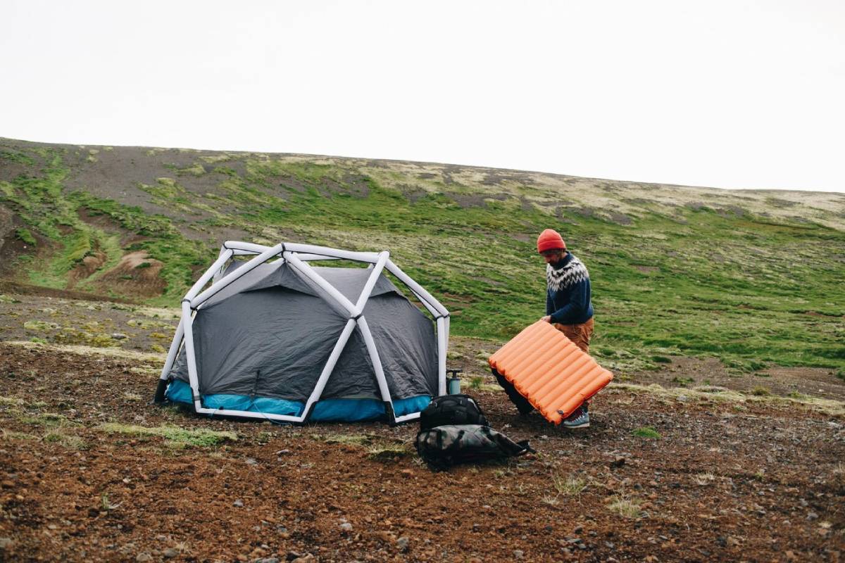 A person in a red hat sets up an orange sleeping pad next to a inflatable geodesic tent on a lush, green hillside. 