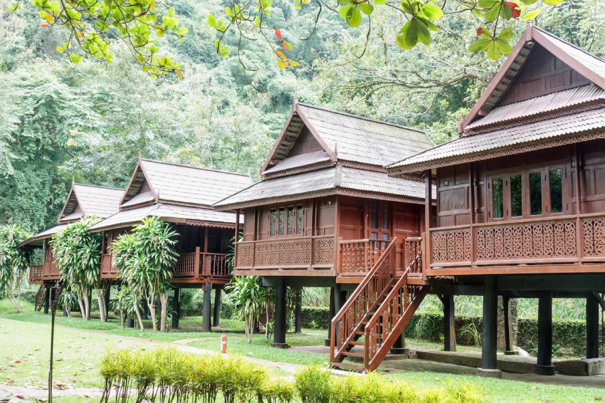 Four traditional wooden cottages on stilts, surrounded by lush greenery, featuring ornate balconies and steeply sloped roofs. 