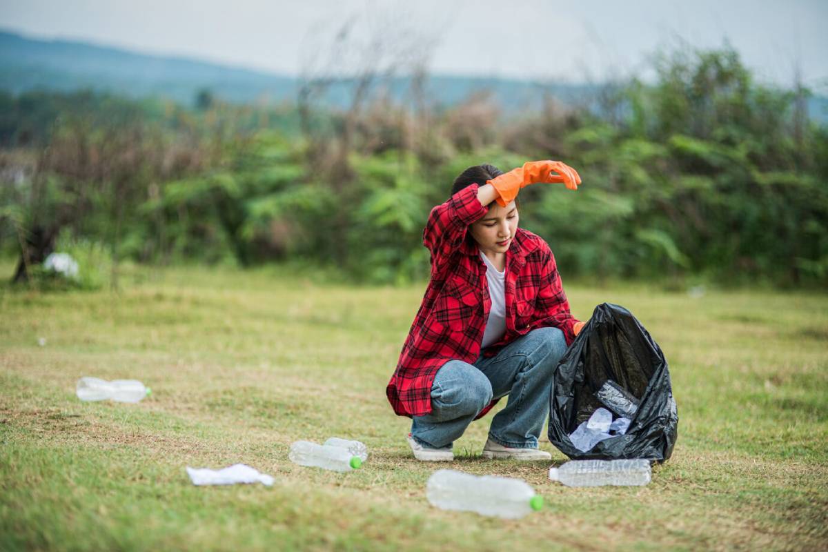 A person in a red plaid shirt and orange gloves crouches on grass, collecting plastic bottles into a black trash bag. 
