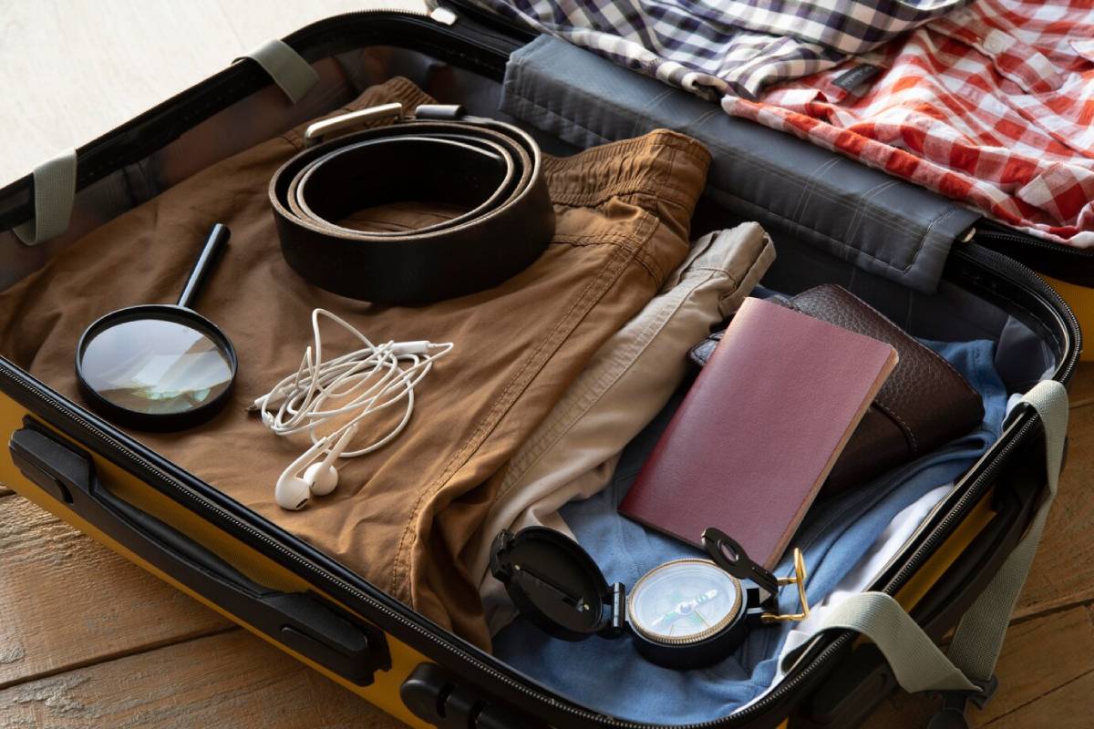 A suitcase packed with clothes, a magnifying glass, a compass, a passport holder, earbuds, and two belts, on a wooden surface. 