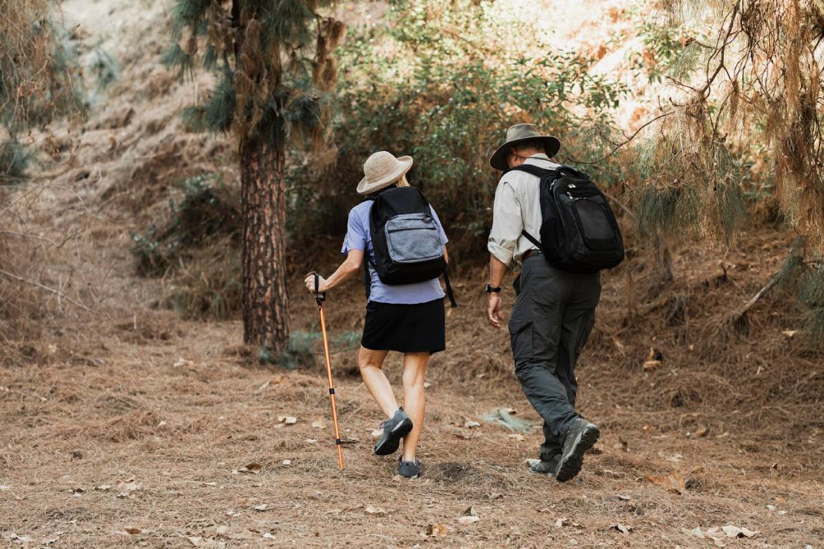  An older couple hiking on a wooded trail, carrying backpacks and using a walking stick, surrounded by trees and dry foliage.