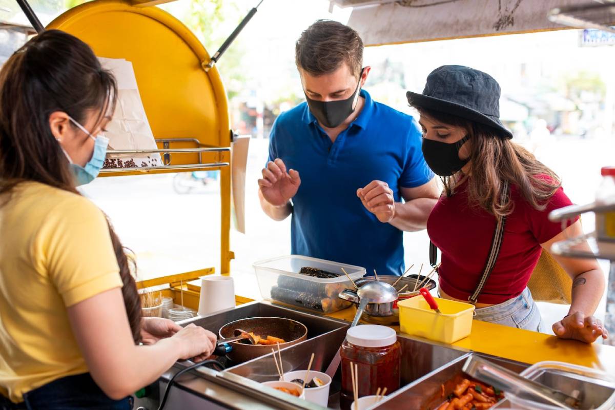 A food vendor serves a couple at a vibrant street food cart, showcasing various dishes and condiments in a lively outdoor setting. 