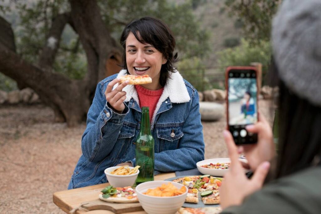 A person enjoys a snack, seated at a table with various dishes and a green bottle, while someone else takes a photo nearby.