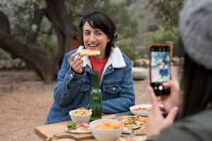 A person enjoys a snack, seated at a table with various dishes and a green bottle, while someone else takes a photo nearby.