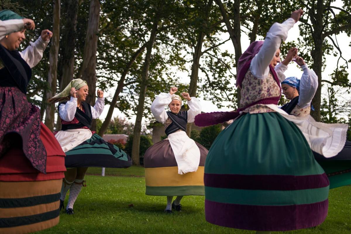 Group of dancers in vibrant, traditional costumes joyfully performing in a lush, green garden, surrounded by tall trees. 