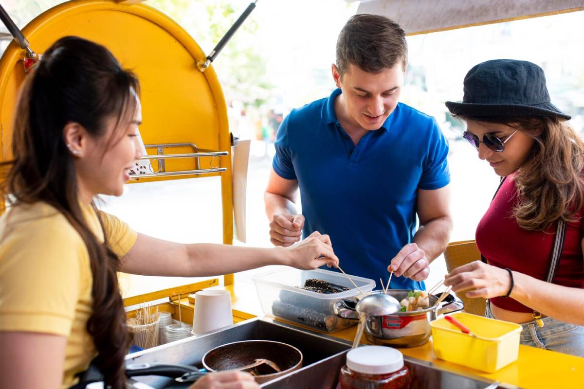 A food vendor serves customers at a vibrant yellow food cart, where two patrons select food items with chopsticks. 