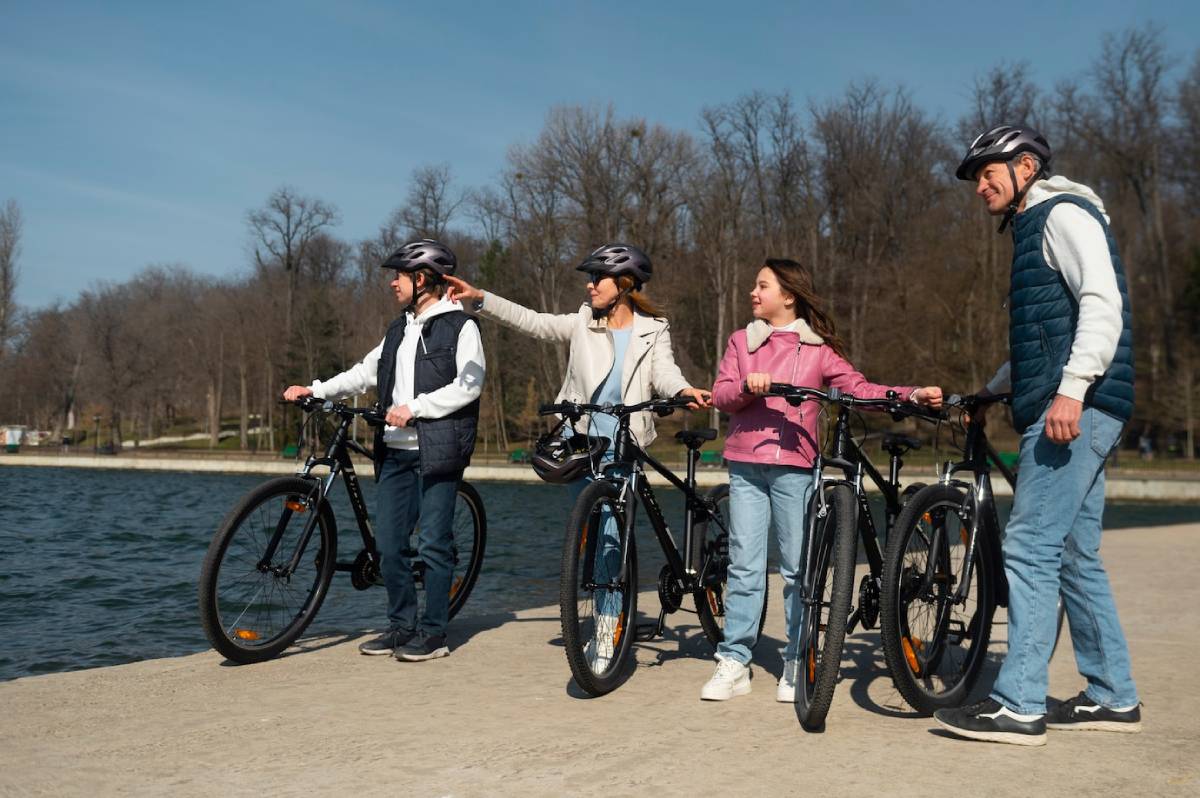 A group of four cyclists stands by a tranquil lake, enjoying a sunny day, with their bikes nearby and trees in the background. 