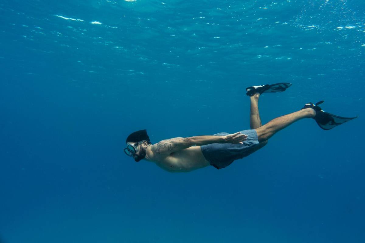  A man with a tattoo swims gracefully underwater, wearing fins and a mask, surrounded by clear blue ocean.