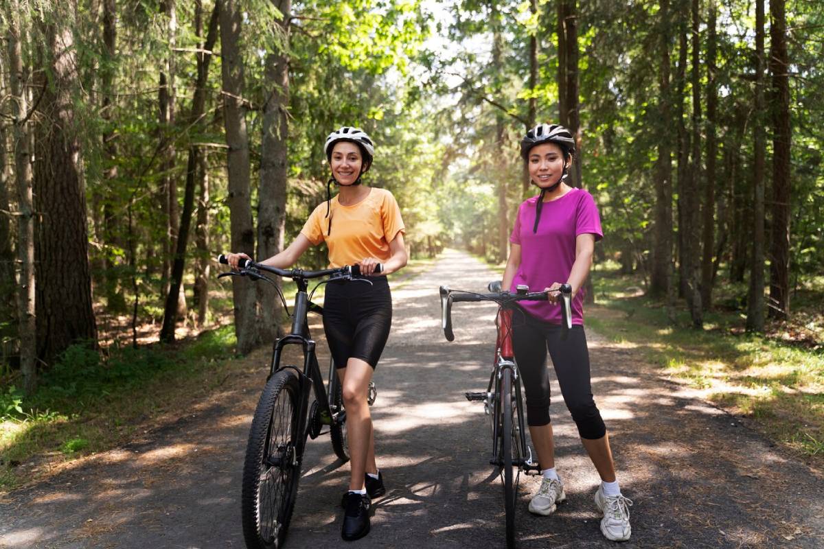  Two young women stand with bicycles on a forested pathway, surrounded by tall trees and dappled sunlight.