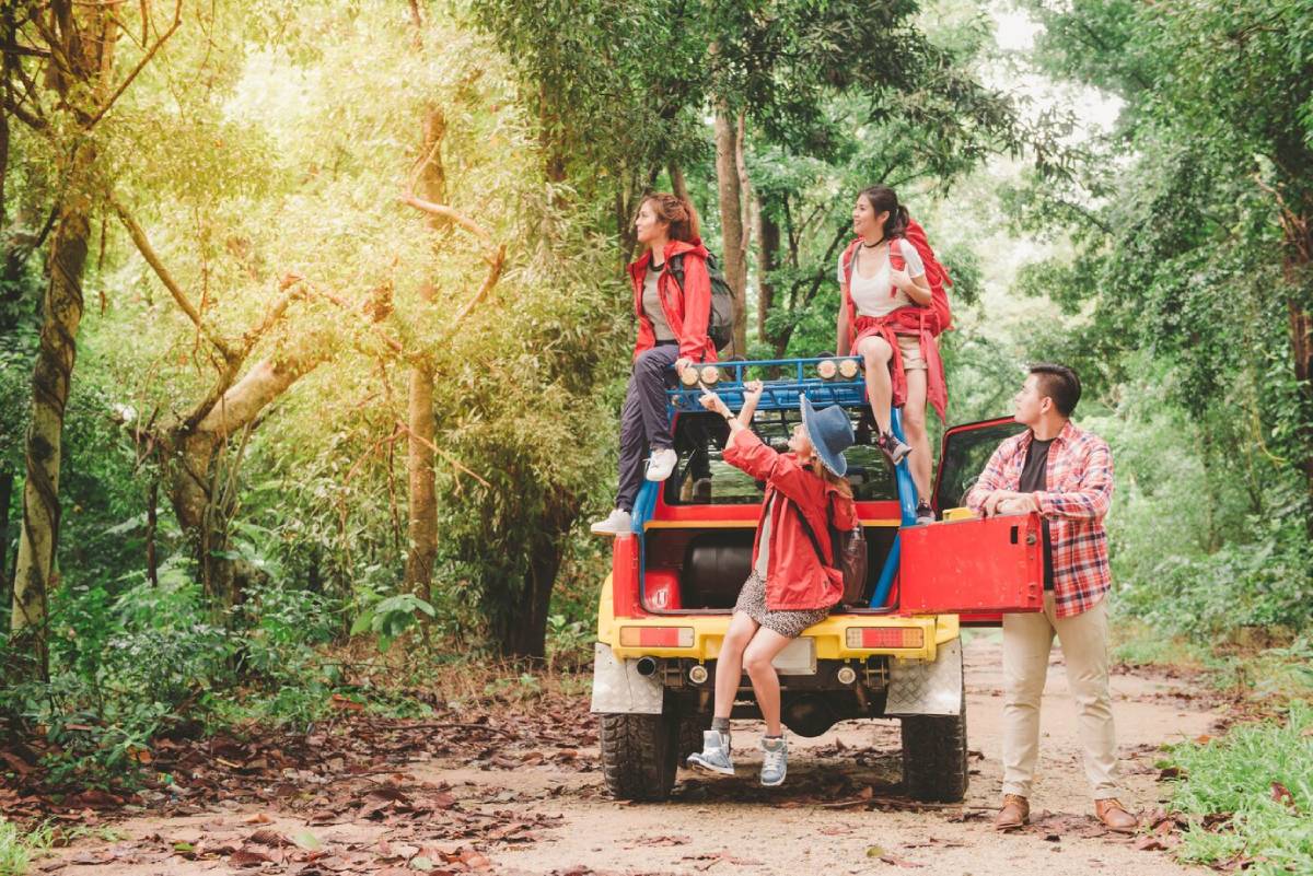  A group of four friends in red jackets enjoying an adventure on a lush forest trail, with a colorful jeep parked nearby.