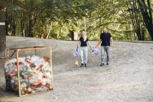 A couple walks on a gravel path in a green park, holding trash bags and tools for cleaning up litter.