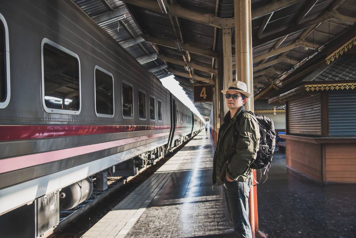  A traveler in a green jacket and hat stands at a train station, with a train parked beside him and wooden buildings in the background.