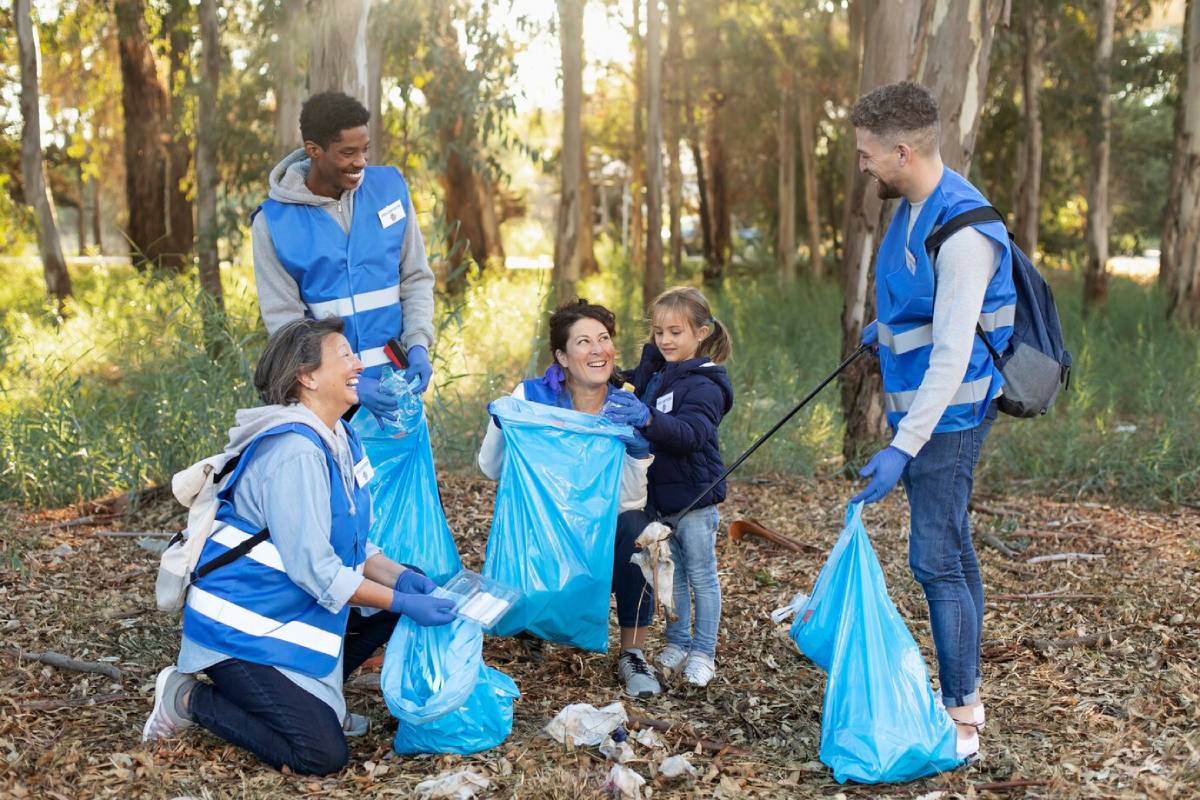  A group of volunteers in blue vests clean up trash in a sunlit forest, holding blue bags and engaging with each other actively.
