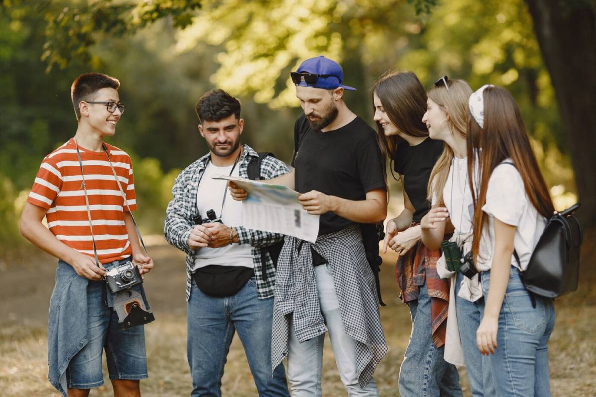 A group of six people in casual clothing gathers outdoors, analyzing a map while surrounded by lush greenery. 