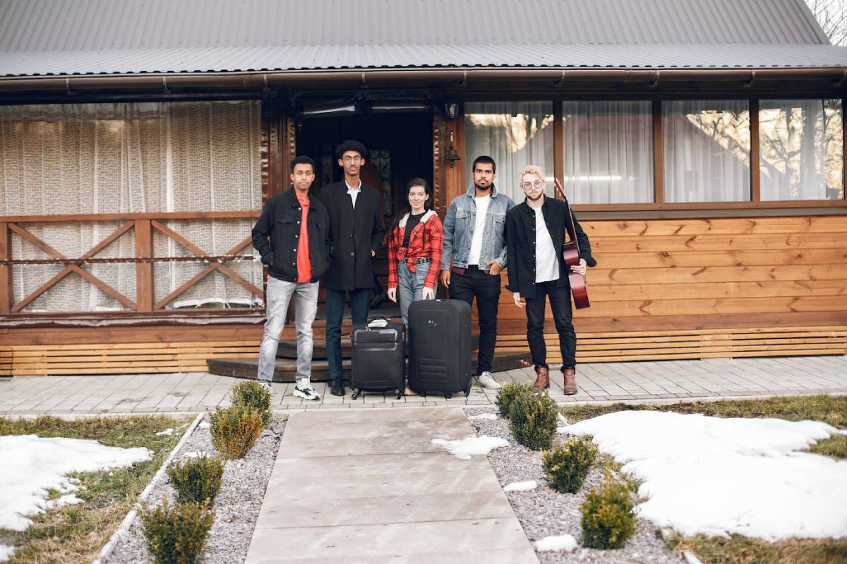  A group of five friends stands in front of a wooden cabin, with luggage and a guitar, amidst a snowy landscape.