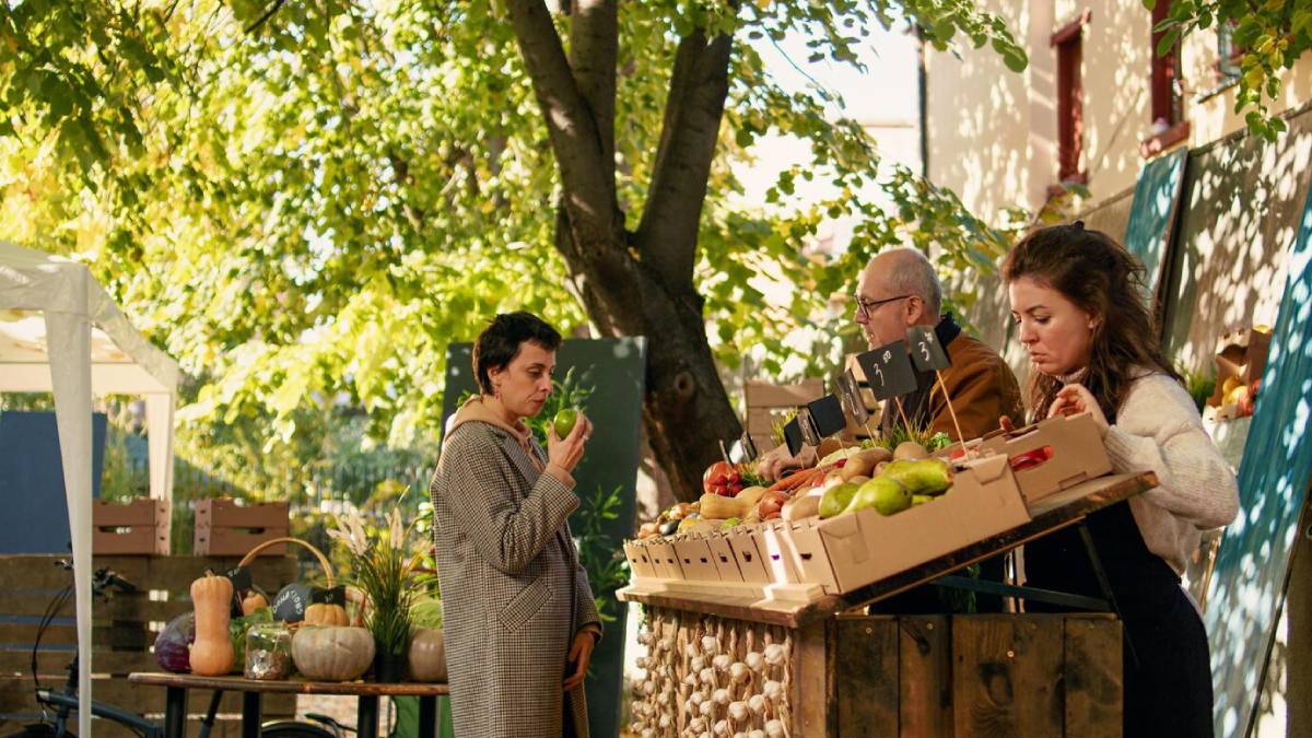 A lively outdoor market scene with a variety of fresh fruits and vegetables displayed on wooden tables, surrounded by greenery. 