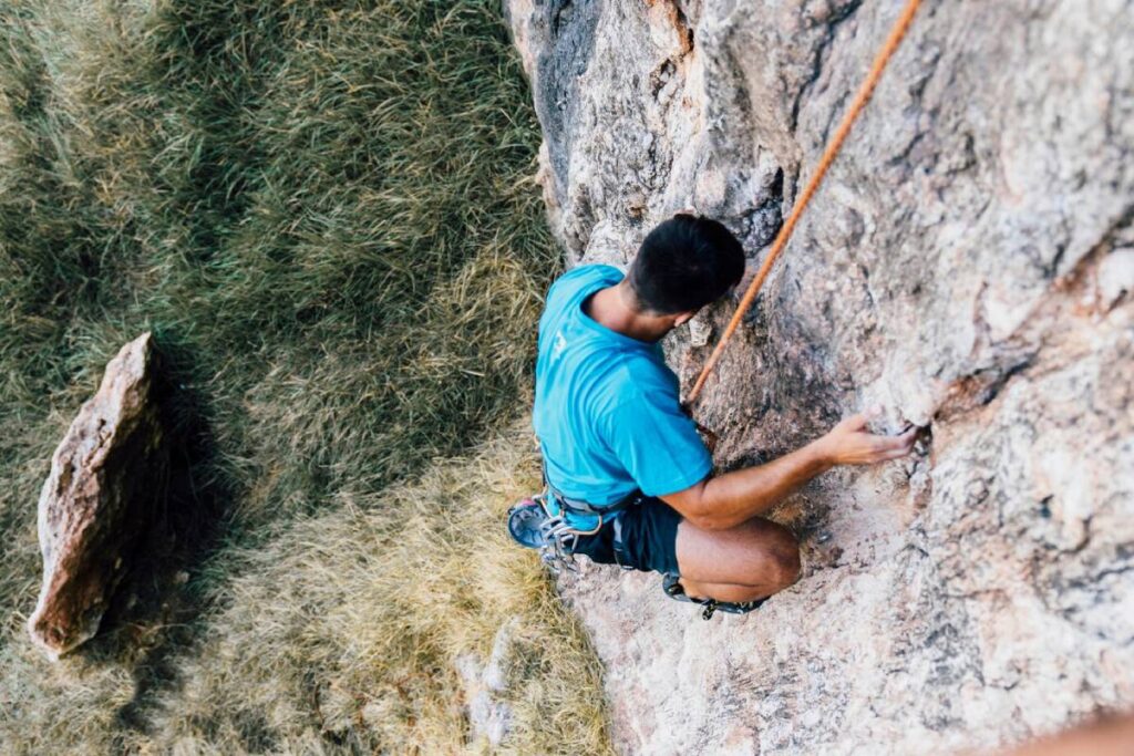 A climber in a blue shirt ascends a rocky cliff, using a harness and rope, surrounded by dry grass and stones below.